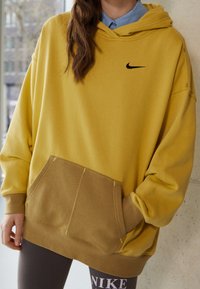 Felpa gialla con logo Nike nero, dotata di tasca frontale e parte inferiore marrone a contrasto. Tessuto morbido con design rilassato.
