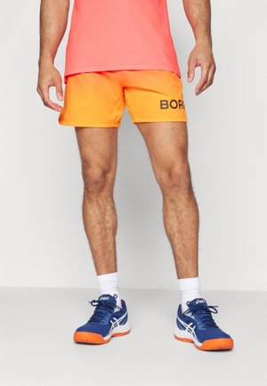 Homme portant un short orange vif avec le texte noir « BORG », un t-shirt corail, des chaussettes blanches et des baskets Asics bleues avec des semelles orange, debout sur un sol gris.