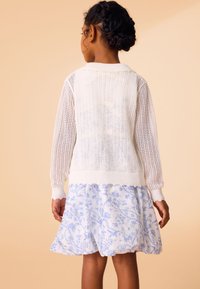 Jauna mergaitė su pintinėmis kasomis, dėvinti baltą mezginį megztinį ir mėlynai baltą gėlėtą sijoną, stovinti priešais smėlio spalvos foną.