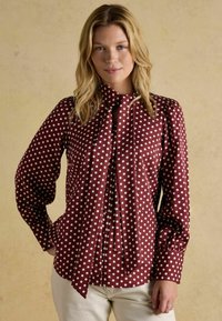 Maroon polka dot blouse with long sleeves, bow tie collar, and button-down front. Smooth fabric and white dots create a classic pattern.
