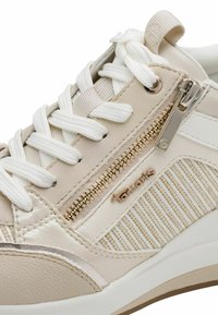 Baskets beige avec accents texturés blancs, dotées d'une fermeture éclair latérale, de ferrures dorées et de lacets plats blancs. Design léger avec des détails subtils.