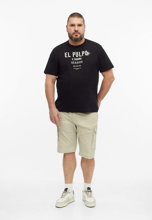 Mężczyzna z brodą i związanymi włosami, ubrany w czarny t-shirt z nadrukiem, beżowe szorty typu cargo, białe skarpetki i białe trampki, stoi z jedną ręką za plecami.
