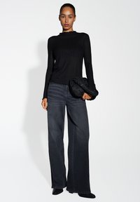 Zwarte geribbelde lange mouw top met een scalloped halslijn, gecombineerd met wijde donkere spijkerbroeken. Model houdt een getextureerde zwarte clutch vast.