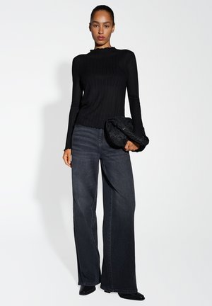 Top lungo nero a costine con scollatura ondulata, abbinato a jeans in denim scuro a gamba larga. Il modello tiene in mano una borsa a tracolla nera texturizzata.