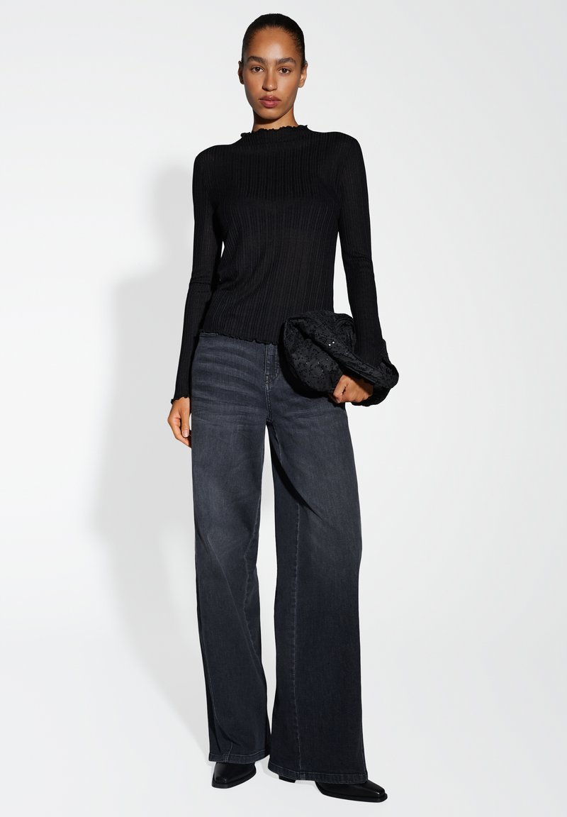 Zwarte geribbelde lange mouw top met een scalloped halslijn, gecombineerd met wijde donkere spijkerbroeken. Model houdt een getextureerde zwarte clutch vast.