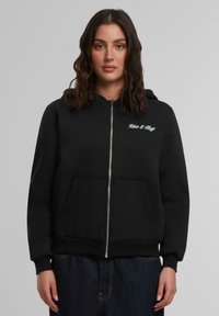Zwarte rits hoodie, gemaakt van een katoenmengsel, met een voorzak en de zin "Rise & Stay" in witte letters op de linker borst.