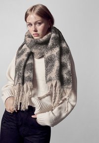 Ung kvinde iført et stort grå- og beige fransesjal, en elfenbensfarvet sweater og sorte bukser, stående med hænderne i lommerne mod en ensfarvet baggrund.