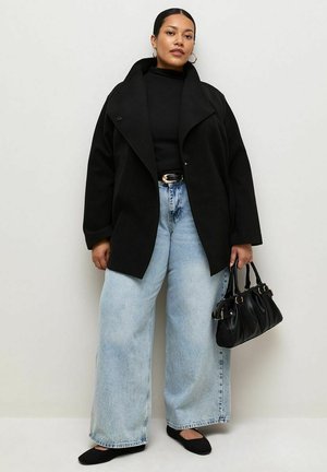 Mujer con abrigo negro, camiseta negra, vaqueros anchos azul claro, bailarinas negras y sosteniendo un bolso negro sobre un fondo claro y liso.