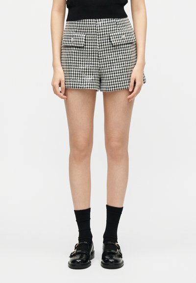 Γυναίκα με σορτς μαύρο-άσπρο ψαροκόκκαλο με μπροστινές τσέπες με καπάκι, μαύρες κάλτσες και μαύρα δερμάτινα παπούτσια με αγκράφα, στέκεται μπροστά σε λευκό φόντο.