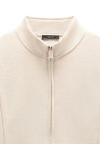 Beżowy top z wysokim kołnierzem zapinany na zamek, z widocznymi przeszyciami i metką „Parfois Collection” wewnątrz kołnierza, suwak również z logo „Parfois”.