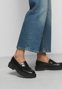 Des mocassins en cuir noir avec une semelle épaisse et un détail à franges, associés à un pantalon en denim large bleu clair, montrant un ourlet brut.