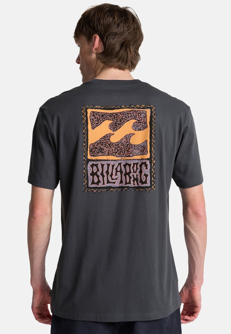 Mann trägt ein dunkelgraues T-Shirt mit orange-lila Billabong-Logo, das ein stilisiertes Wellenmuster auf der Rückseite zeigt.