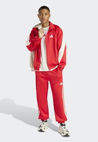 Mladic u crvenom i bijelom Adidas trenirci s bijelom majicom, drži jaknu preko ramena, stoji uz jednostavnu pozadinu.