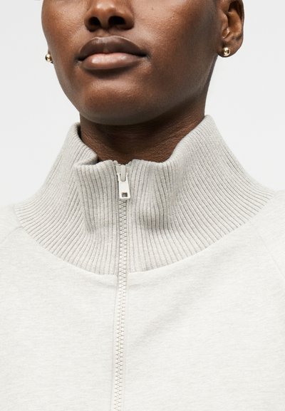 Κοντινό ενός ατόμου που φοράει ανοιχτό γκρι πουλόβερ με φερμουάρ, ριμπ ύφασμα στον ψηλό γιακά και χρυσά σκουλαρίκια με καρφίτσα, κλειστά χείλη, ουδέτερη έκφραση.