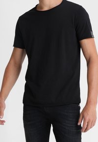 Sort t-shirt med rund hals, korte ærmer og en diskret logo detalje på venstre ærme; lavet af blødt, strækbart stof.