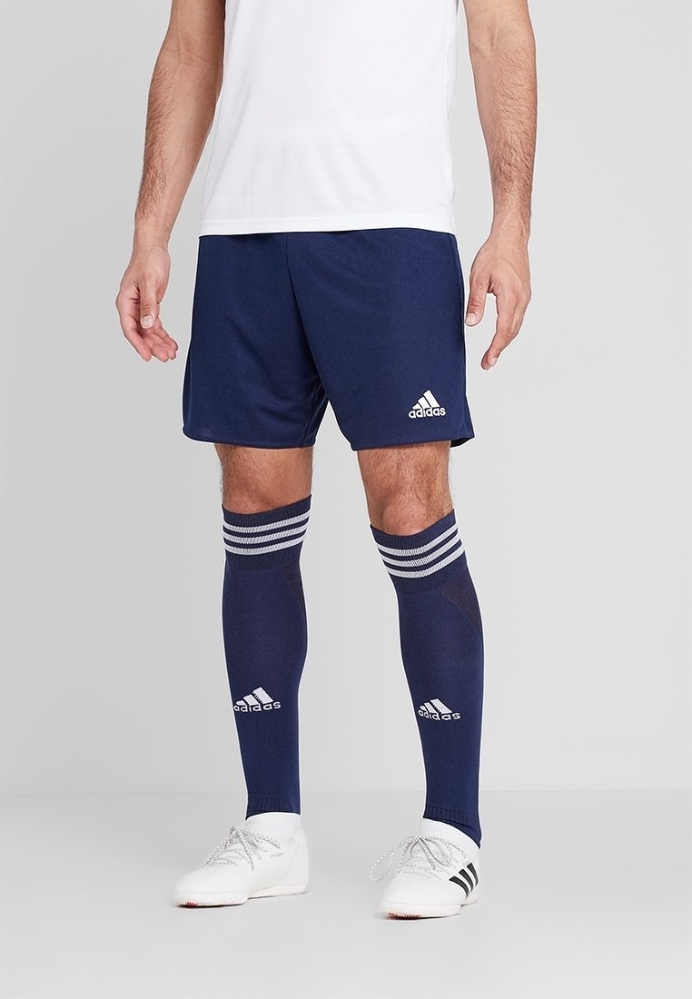 Muž ve tmavě modrých sportovních šortkách Adidas, odpovídajících ponožkách po kolena s pruhy, bílých kopačkách Adidas a bílém tričku na hladkém šedém pozadí.