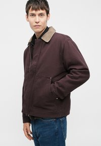 Vinsdækket zip-jakke med kordfløjlskrave, forlommer og struktureret stof. Sammen med blå jeans, præsenteret mod en ensfarvet baggrund.