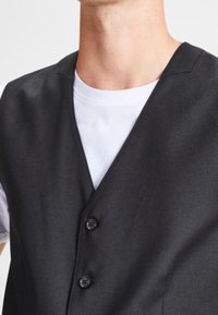 Tummanharmaa liivi, jossa on V-pääntie, kolme nappia ja sileä rakenne, kerrostettuna valkoisen T-paidan päälle. Yksinkertainen, räätälöity muotoilu.