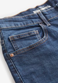 Tummansiniset farkut, joissa on kontrastivärinen oranssi tikkaus ja pieni takatasku. Kankaan pinta on sileä ja siinä on lievää kulumista.