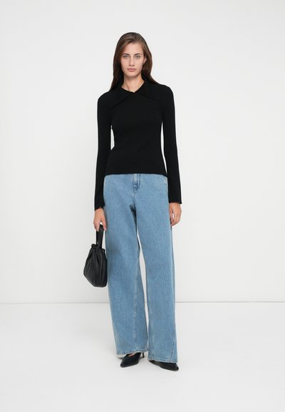 Svart langermet topp med bred krave, kombinert med lyseblå vide jeans. Modellen holder en svart poseveske og har på seg svarte spisse sko.