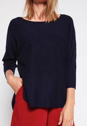 Femme portant un haut ample à manches longues bleu marine et un pantalon rouge taille haute, une main dans la poche, fond neutre.