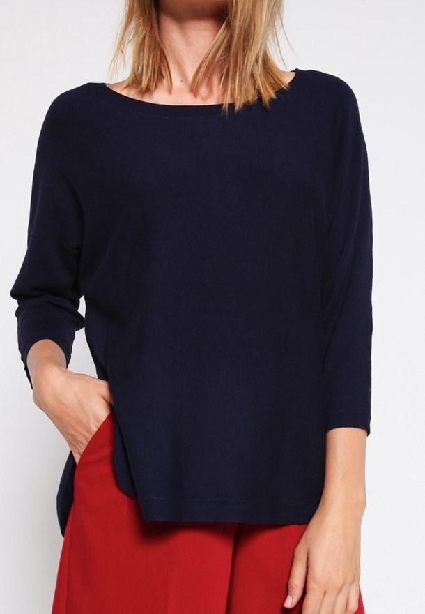 Femme portant un haut ample à manches longues bleu marine et un pantalon rouge taille haute, une main dans la poche, fond neutre.