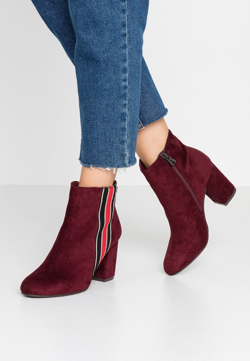 Bottes en suède bordeaux montant à la cheville, à talons blocs, avec une fermeture éclair latérale et un détail rayé rouge, blanc et noir, portées avec un jean bleu raccourci.