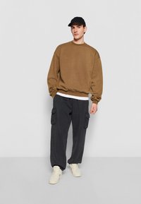 Brun, oversized sweatshirt, tapered svarta cargobyxor med sidofickor, och vita sneakers, framhävt av en svart keps.