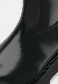 Schwarzer, glänzender Knöchelboot mit einem schlanken Design, glatter Textur und dezenten Nähakzenten entlang der oberen Naht und Sohle.