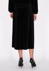 Sort velour midi-nederdel med en glat tekstur, der har en blød silhuet. Kombineret med sorte højhælede sko, der har et enkelt design.