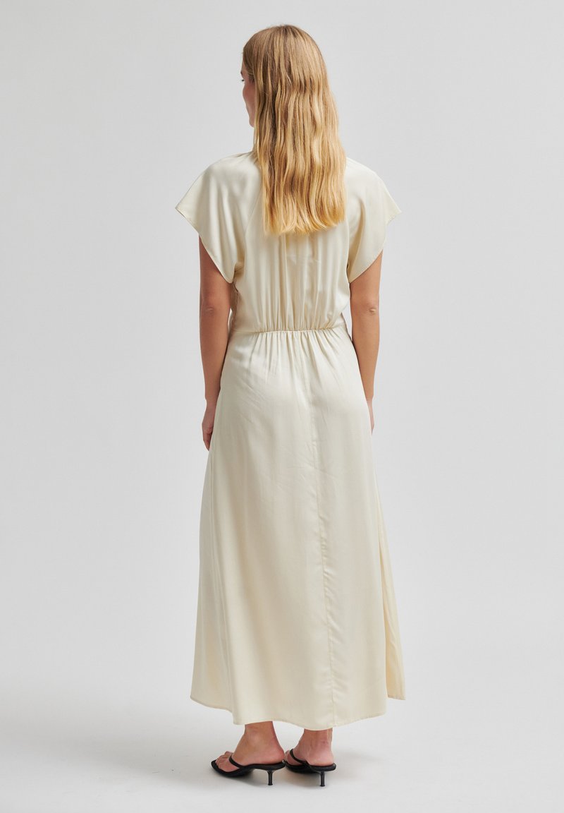 Femme aux longs cheveux blonds portant une robe crème à manches courtes et longueur cheville, ainsi que des sandales noires à talons, debout de dos.