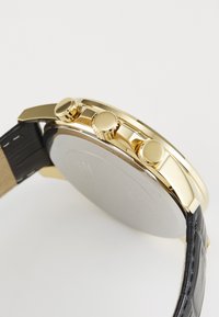 Montre en métal doré avec un bracelet en cuir noir, équipée de trois boutons de couronne et d'un dos en acier inoxydable lisse.