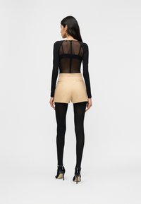 Γυναίκα που φοράει διαφανές μαύρο τοπ με μακριά μανίκια, μπεζ σορτς, μαύρες καλσόν και μαύρα παπούτσια με ψηλό τακούνι, όρθια, γυρισμένη προς τα πίσω σε απλό φόντο.