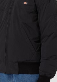 Veste bomber Dickies noire zippée avec poignets et taille côtelés, portée par-dessus un jean bleu.