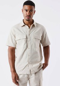 Camisa de botón de manga corta en color beige, hecha de tela texturizada, que presenta dos bolsillos en la parte frontal, un collar puntiagudo y un dobladillo recto.