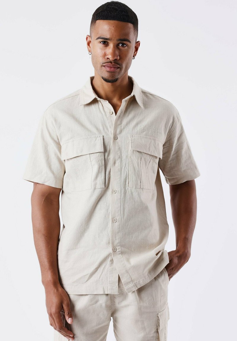 Camisa de botón de manga corta en color beige, hecha de tela texturizada, que presenta dos bolsillos en la parte frontal, un collar puntiagudo y un dobladillo recto.