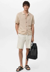 Beige short-sleeve knit shirt with button front, cream shorts, black backpack, and black sandals. Simple, lightweight design and neutral colors.
