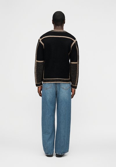 Άτομο που φοράει μαύρο πουλόβερ με μπεζ ραφές και μπλε τζιν παντελόνι με φαρδιές μπατζάκια, στέκεται γυρισμένο με την πλάτη προς το μέρος μας μπροστά σε απλό λευκό φόντο.