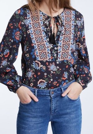Kvinde iført en sort blomstret bluse med broderede lodrette paneler og bindebånd ved halsen, kombineret med blå cowboybukser, hænderne i lommerne.