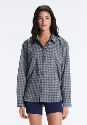 Hosszú sötét hajú nő, aki szürke és fekete kockás hosszú ujjú inget és tengerészkék rövidnadrágot visel, egy egyszínű háttér előtt áll.