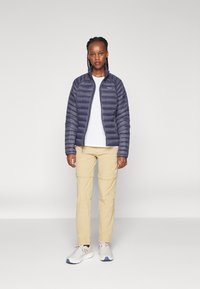 Tamsiai mėlynas pūkinis švarkas su užtrauktuku priekyje ir stovinčiu apykakle, derinamas su smėlio spalvos kelnėmis ir pilkiais sportiniais batais. Matomas lygus audinys ir kvadratinis dizainas.