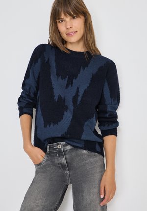 Vrouw in een marineblauwe gebreide trui met opgerolde mouwen, gecombineerd met vervaagde grijze jeans, staand tegen een effen witte achtergrond.