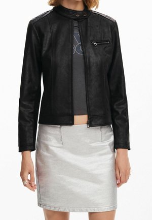 Kobieta w czarnej skórzanej kurtce na suwak, ciemnym wzorzystym topie i błyszczącej srebrnej mini spódnicy, stojąca na białym tle.