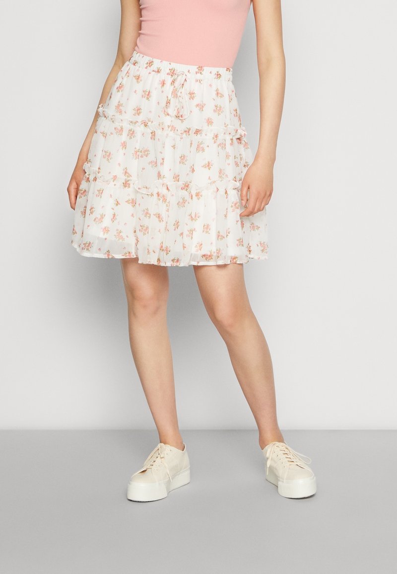 Femme portant une jupe blanche à volants fleurie, un haut sans manches rose clair et des baskets compensées écrues, debout sur un sol gris.