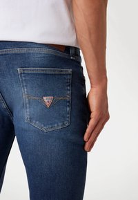 Denim jeans in dark blue, featuring a stitched back pocket with a red and white triangle logo. Smooth texture with a fitted shape.