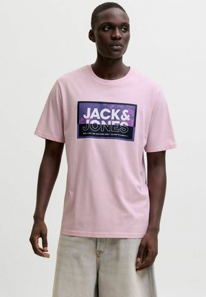 Young man wearing a light pink T-shirt with a purple "JACK & JONES" logo block and light gray jeans, standing against a plain background.