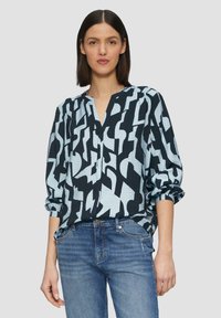 Blouse à manches longues avec un motif géométrique audacieux noir et bleu clair, col à boutonnière et coupe décontractée. Associée à un jean bleu.