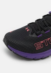 La chaussure de sport noire et violette présente un dessus en mesh, un logo Under Armour rouge et une inscription "ST" en rouge audacieux sur le côté, avec des lacets noirs.