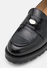Mocassin en cuir noir à finition brillante, avec un détail en métal argenté portant l'inscription "BOSS" et une semelle en caoutchouc texturée.