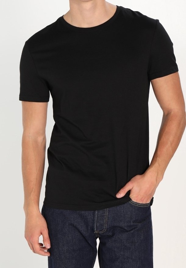 Homme portant un t-shirt noir uni à manches courtes et un jean bleu foncé, avec une main dans la poche, sur un fond blanc.